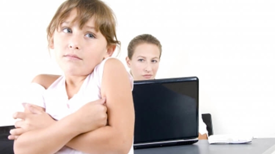 A young girl is shrugging whie a woman behind her works on a black laptop.
