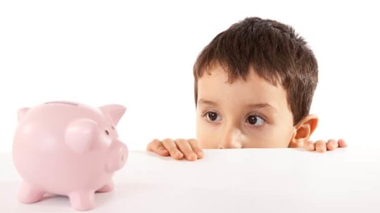A young boy peeks over the top of a table at a piggy bank.
