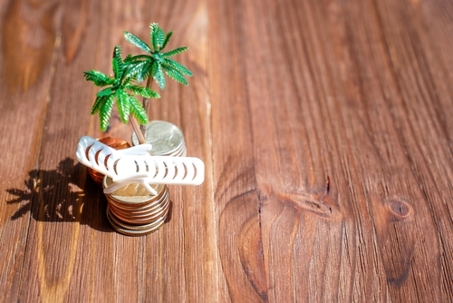 Coins stacks and a toy beach lounger under miniature palm trees set on a dark wooden background. Offshore financial centers and tax avoidance concept.
